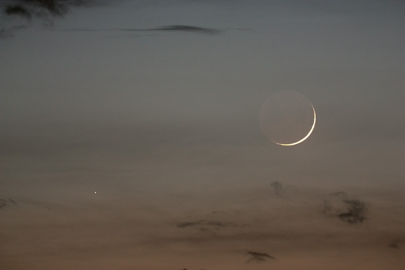 Vor einem noch hellen Abendhimmel in der Dämmerung leuchtet rechts eine zarte Mondsichel, die Nachtseite des Mondes ist als blase Scheibe erkennbar. Links leuchtet Merkur als kleiner Lichtpunkt.