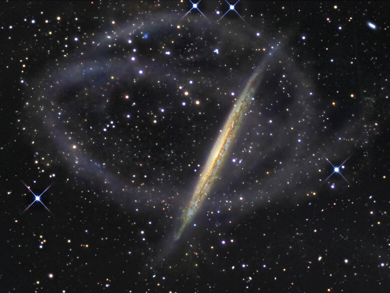 Das Bild zeigt eine verkrümmte, von der Seite sichtbare Galaxie, die von Sternströmen umschlungen ist.