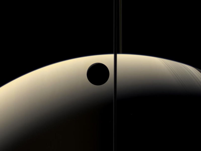 Vor der riesigen Sichel Saturns, die waagrecht im Bild liegt und rechts und links abgeschnitten ist, zeichnen sich der Mond Rhe als schwarze Scheibe und das Ringsystem als schwarze Linie ab.
