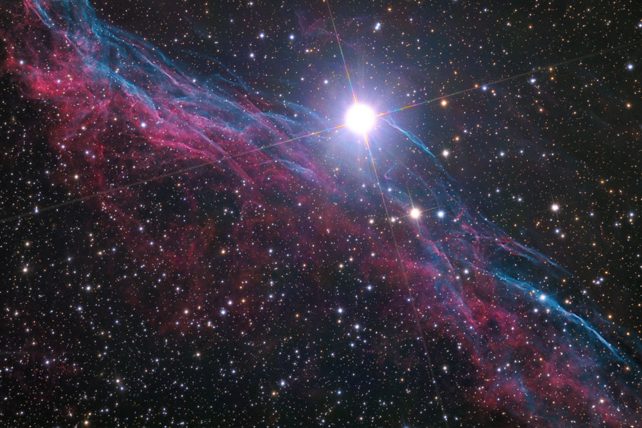 Unter einem hellen Stern mit Lichtkreuz verläuft diagonal ein purpur-bläulicher Nebel aus vielen Fasern. 