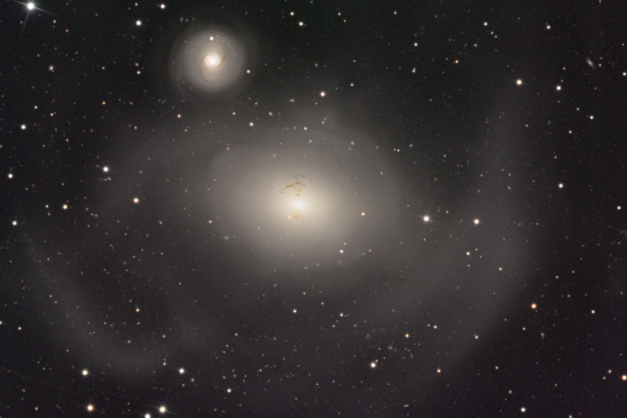 Mitten im Bild leuchtet eine verschwommene elliptische Galaxie, links darüber eine kleinere, ebenfalls verschwommene Galaxie mit leichten Strukturen, beide Galaxien haben einen sehr hellen Kern.