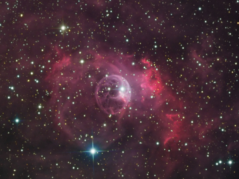 In der Mitte des Sternenfeldes leuchtet ein rosaroter Nebel, in dessen Mitte eine blasenartige Struktur mit einem hellen Stern rechts oben erkennbar ist.