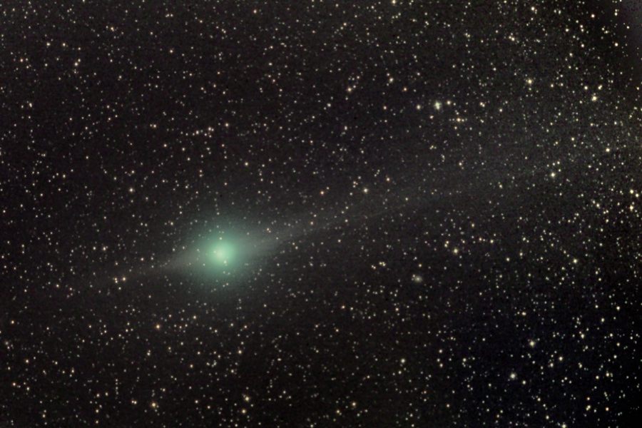 Vor einem Hintergrund mit Sternen leuchtet ein grünlicher Komet, dessen Schweif nach rechts oben verläuft und nur schwer erkennbar ist.