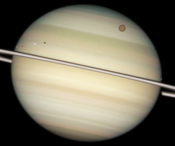 Saturn ist bildfüllend dargestellt, von den Ringen ist nur ein schmales Band zu sehen, sie sind links und rechts abgeschnitten, oben sind einige Monde zu sehen, teils mit Schatten auf dem Planeten.