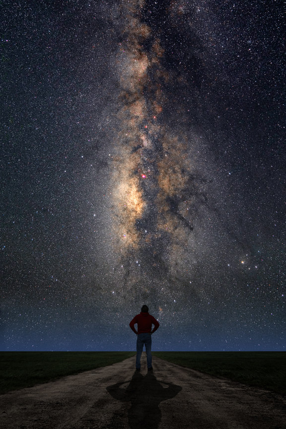 Auf einer Ebene steht ein Mensch, hinter ihm steigt die Milchstraße senkrecht vom Horizont auf.