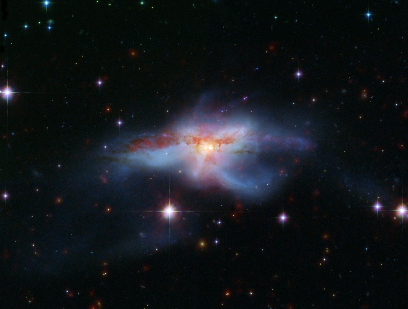 Die Galaxie in der Mitte wirkt wie ein verschwommener, blauer Nebel mit einigen milchigen Ausläufern. Quer über das gelb leuchtende Zentrum verläuft ein rotes gefasertes Staubband.