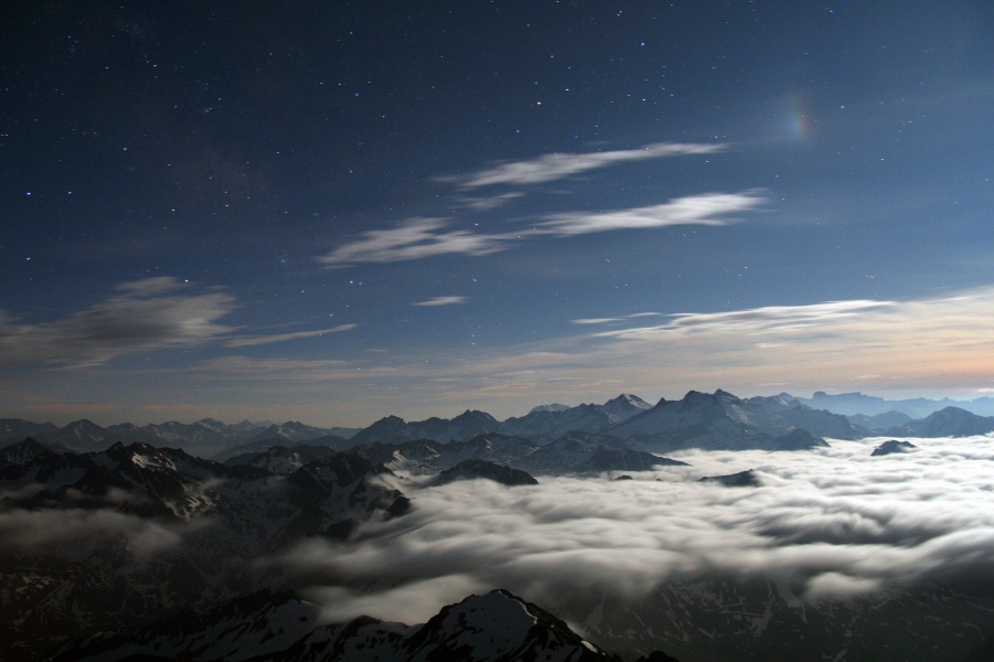 Eine Landschaft mit Bergen und Wolken, darüber ein blauer Himmel mit einigen Wolken, links oben ist ein kleiner regenbogenfarbiger Nebenmond.