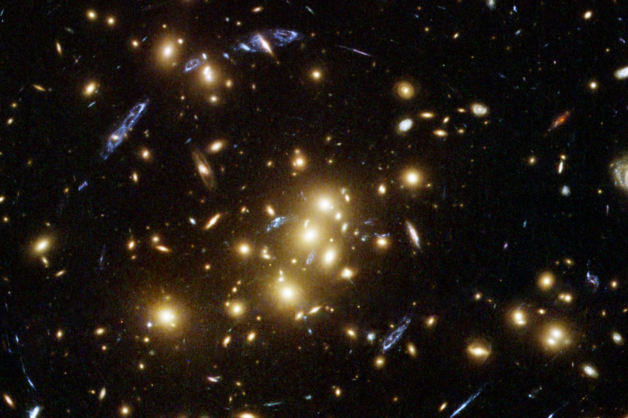 Im Bild sind die Galaxien eines Galaxienhaufens abgebildet, dazwischen sind verzerrte weiter entfernte Galaxien verteilt.