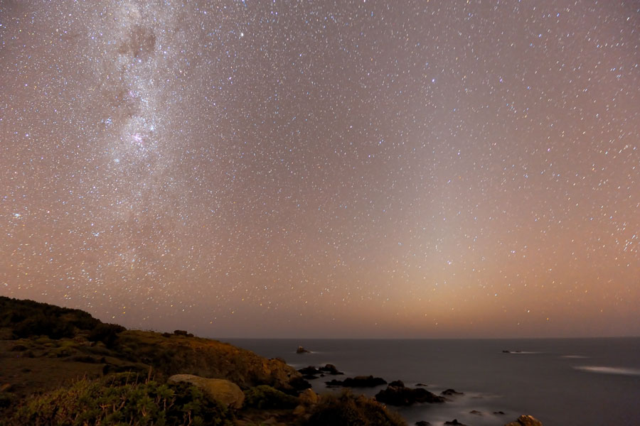 Hinter einem felsigen Strand und dem Meer steigt links die Milchstraße auf, rechts neben der Mitte ist ein zarter Lichtstreifen, das Zodiakallicht, zu sehen.
