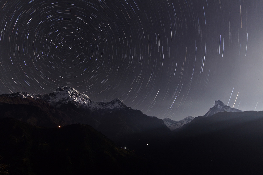 Über einem Panorama mit Berggipfeln sind die Strichspuren von Sternen am Nachthimmel zu sehen.
