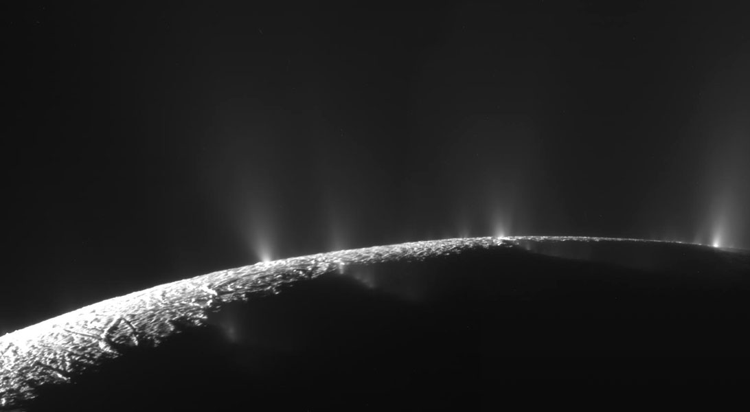 Das Bild zeigt das obere Ende des Mondes Enceladus, links ist ein schmaler, nach rechts spitz zulaufender Streifen von der Sonne beleuchtet. Oben strömen helle Sprühwolken aus der Oberfläche ins All.
