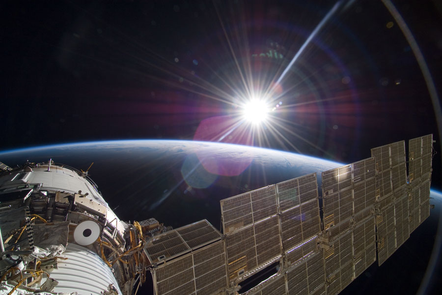 Im Vordergrund ragt unten ein Teil der ISS und ein großes Solarpaneel ins Bild, dahinter geht die gleißende Sonne über der gekrümmten Erde auf.