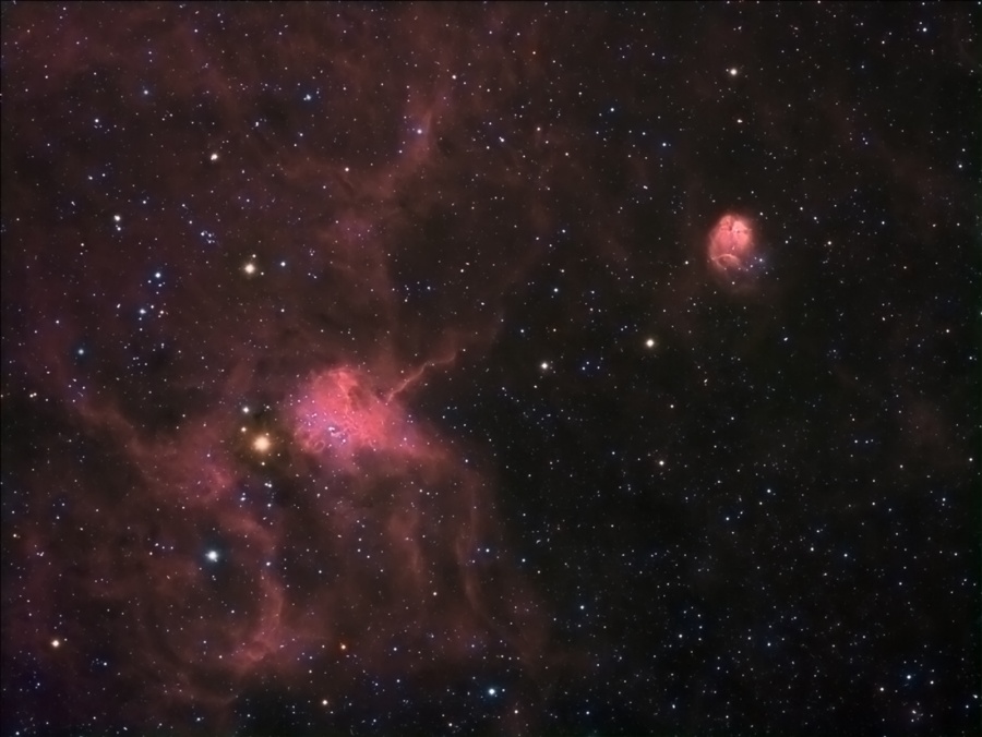 Im Bild leuchten zwei rötliche Nebel, links ein größerer mit Nebeln herum, rechts weiter oben ein kleiner, dazwischen sind Nebel verteilt.