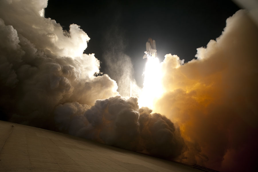 Hinter mächtig aufgetürmten Wolken - links grauweiß, rechts gelblich-dunkel - steigt eine Raumfähre auf einem weißen Feuerstrahl am Nachthimmel auf.