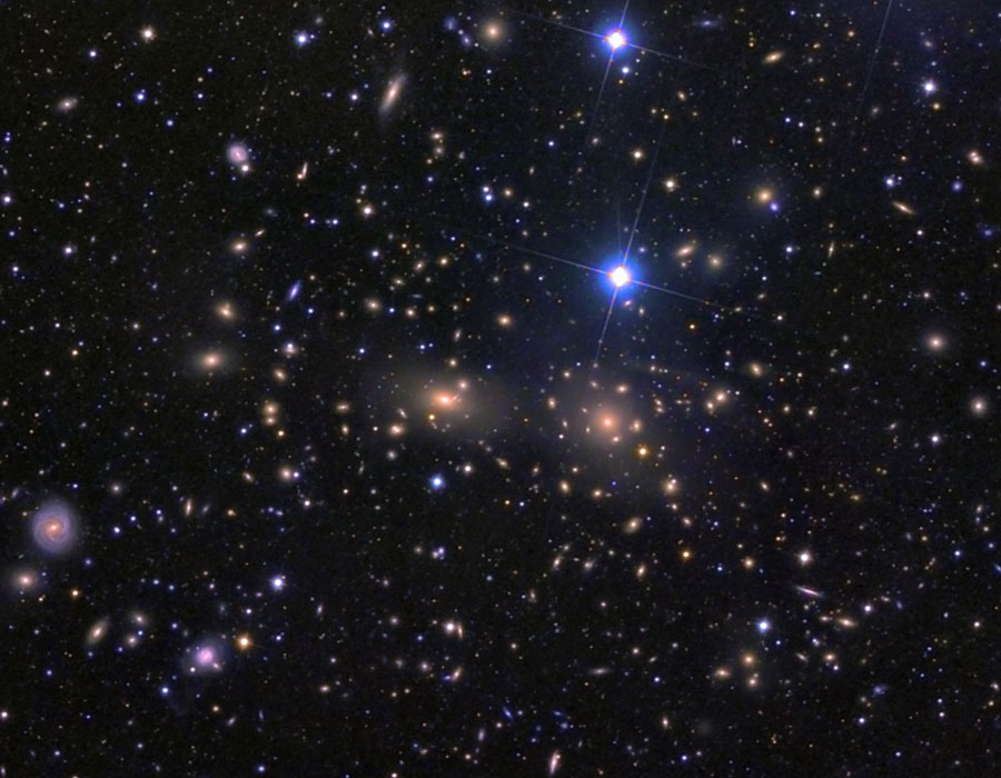 Im Bildfeld sind Galaxien wie Sterne verteilt. In der Mitte leuchten zwei hellorange hervor, oben sind zwei blau leuchtende Sterne, links am Bildrand ist eine markante runde Galaxie.