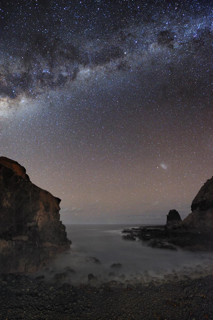Über einem Ufer mit interessanten Felsgebilden leuchtet die Milchstraße mit markanten Dunkelnebeln, rechts unten ist die Kleine Magellansche Wolke.