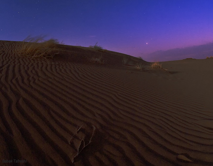 Hinter einer Sanddüne leuchten in der Dämmerung am purpurfarbenen Himmel mehrere Planeten, die entlang der Ekliptik nach rechts aufsteigen.