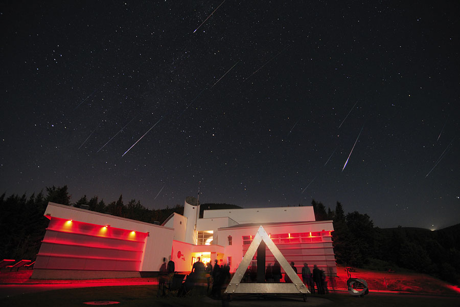 Über einem rot beleuchteten Gebäude leuchten Sterne, und am Himmel blitzen Meteore herab.