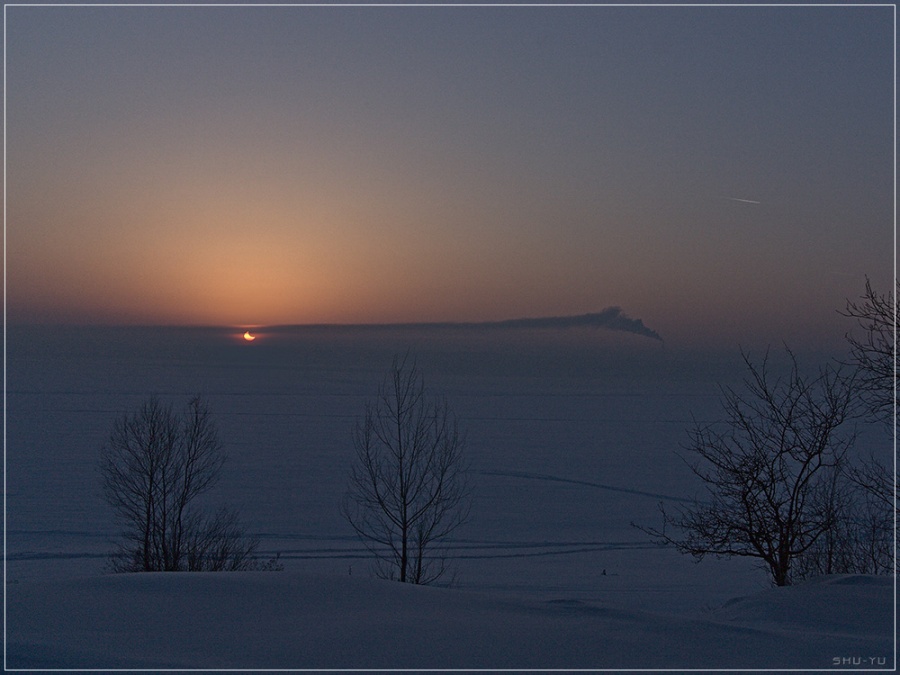 Hinter einer verschneiten Landschaft hängt die partielle Sonnenfinstrnis über dem Horizont, zusammen mit einer Abgasschwade.