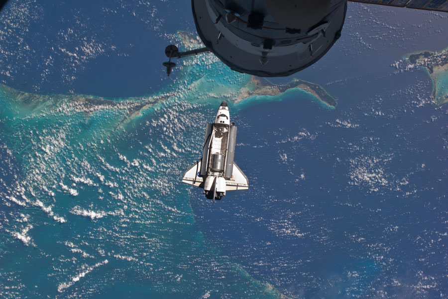 Über einigen Inseln im Meer schwebt die Raumfähre Atlantis unter der Internationalen Raumstation.