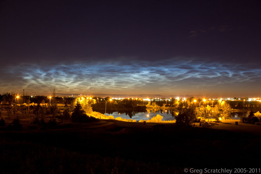 Über einer lichtverschmutzten Stadt mit einem See im Vordergrund breitet sich ein blau leuchtendes Wolkenmuster am Himmel aus.
