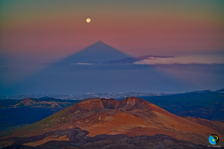 Hinter einem felsigen Vordergrund ragt der Schatten eines Berges bis zum Horizont. Darüber geht der Mond im Abendrot auf.
