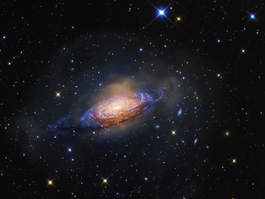 Mitten im Bild ist eine Spiralgalaxie, die etwas unregelmäßig wirkt. In der Mitte ist sie hellgelb, zum Rand hin sind markante Staubbahnen verteilt, ihre leicht derzerrten äußeren Gebiete leuchten boau, und sie ist in Nebel gehüllt.