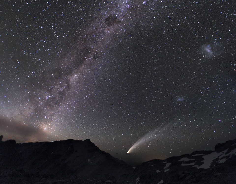 Über einem dunklen Gebirge strahlt ein heller Komet mit gefiedertem Schweif. Von Links steigt die Milchstraße diagonal steil nach oben, rechts sind am sternklaren Himmel die magellanschen Wolken zu sehen.