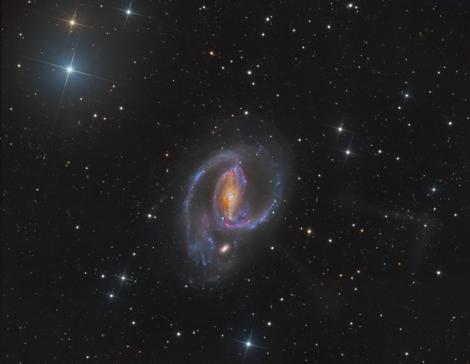 In der Mitte schwebt eine Balkenspiralgalaxie, der Balken in der Mitte leuchet gelblich mit rötlichen Staubbahnen, von diesem gehen zwei gekrümmte ausgeprägte blaue Spiralarme aus. Im Hintergrund sind wenige Sterne.