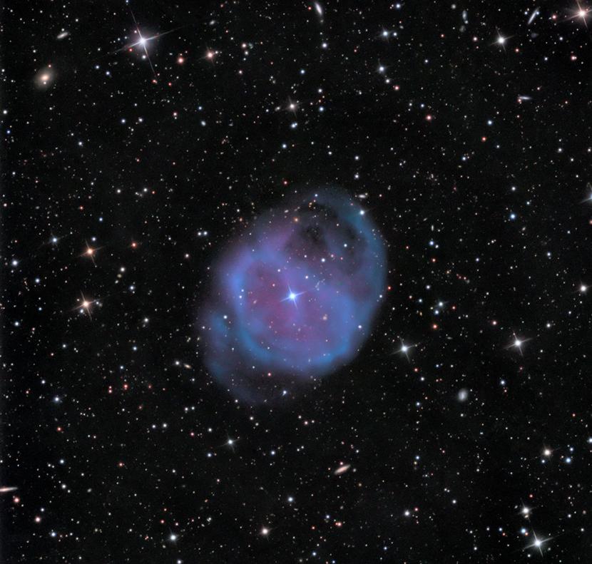 Vor einem sternbestreuselten Hintergrund leuchtet ein blauvioletter Nebel mit einem hellen Stern in der Mitte und einigen schleifenartigen Strukturen.