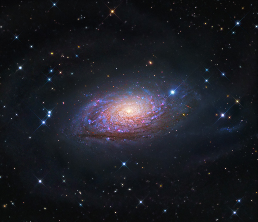 Die Galaxie, die im Bild schwebt, wirkt ein wenig verschwommen. Sie besitzt gleichmäßig gewundene Spiralarme mit vielen Sternhaufen und Sternbildungsregionen. Das Zentrum leuchtet gelblich.