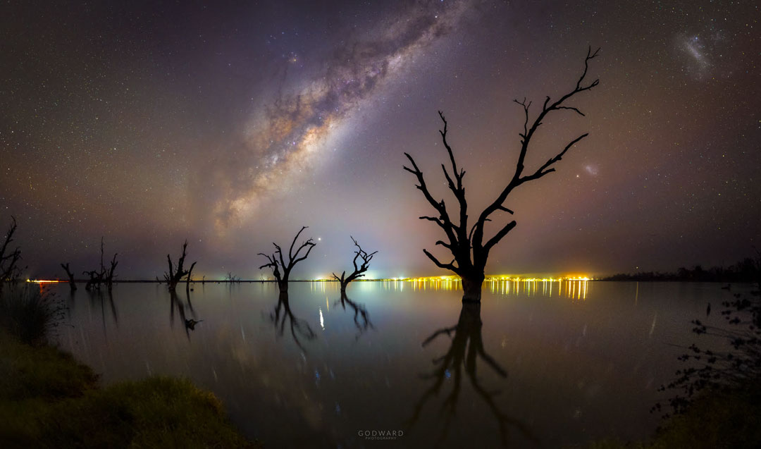 Milchstraße, Planeten, Bäume und Stadtlichter am Ufer von Lake Bonney Riverland in Südaustralien.