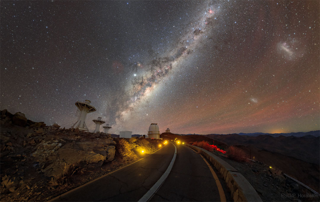Das Bild zeigt die Straße zum La Silla Observatorium in Chile mit einem Teleskop am Horizont und Sternen, Galaxien, Planeten und Nachthimmellicht.