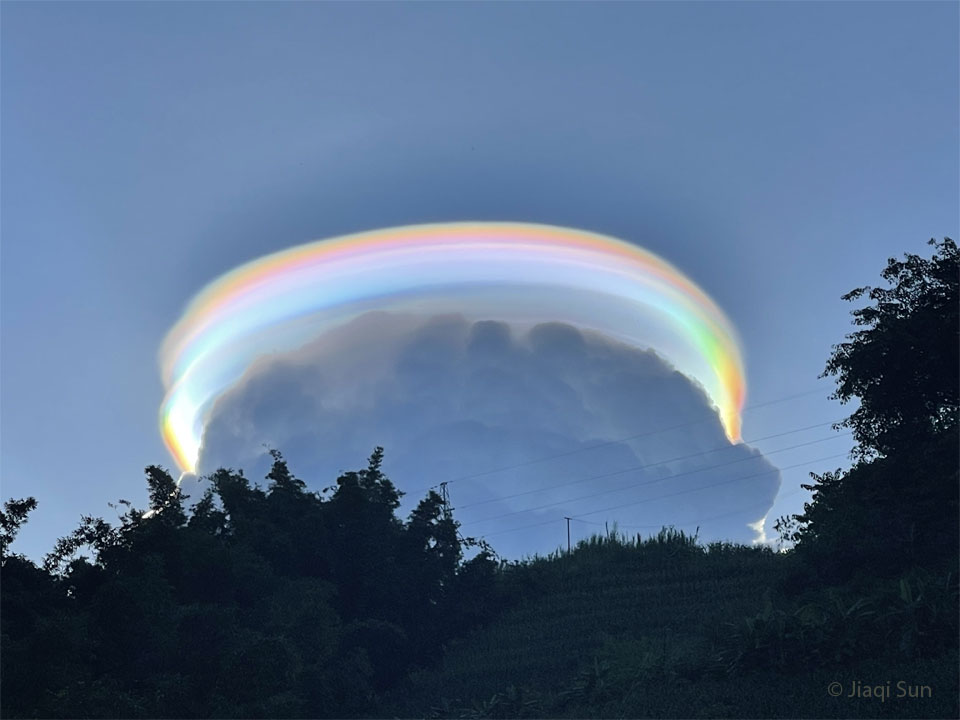 Das Bild zeigt eine in allen Regenbogenfarben schillernde Pileuswolke über einer dunkleren Wolke.