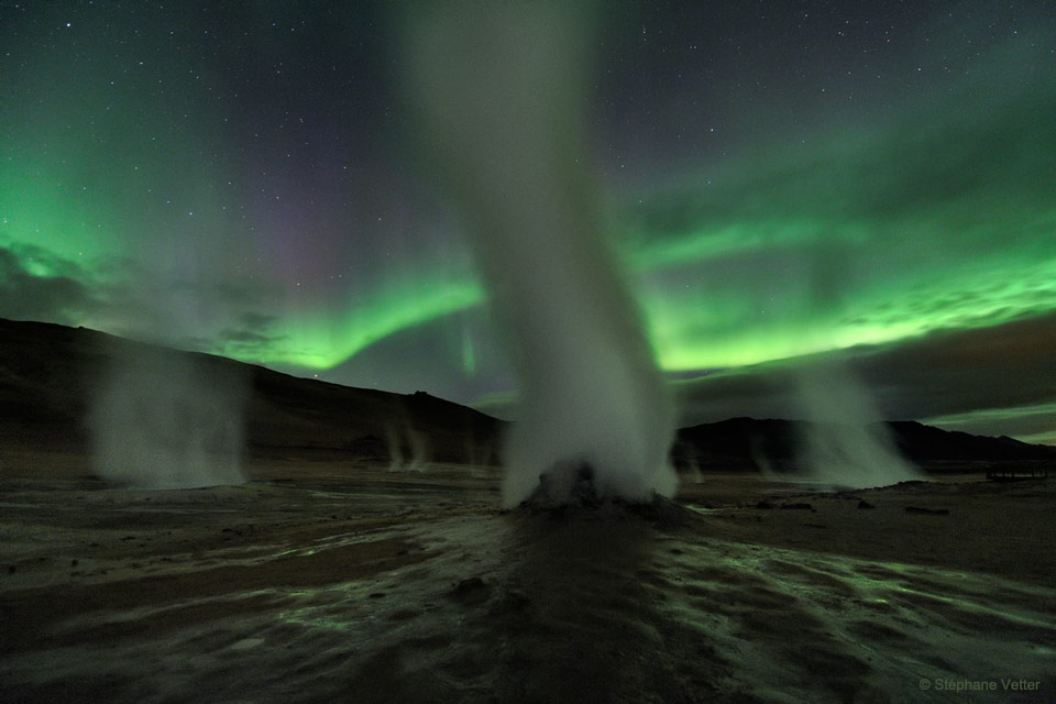 Das Bild zeigt Dampfsäulen, die aus Fumarolen in der isländischen Region Hverir srömen, im Hintergrund leuchtet ein grünliches Polarlicht.