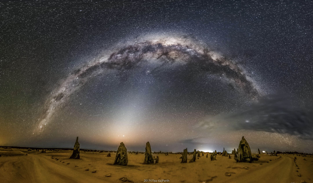 Das Bild zeigt eine Landschaft mit schmalen, dreieckigen Felsen vor einem Nachthimmel, über dem sich das Band der Milchstraße wölbt. Das Land ist durch ein helles, diffuses Zodiakallicht mit dem Himmel verbunden.