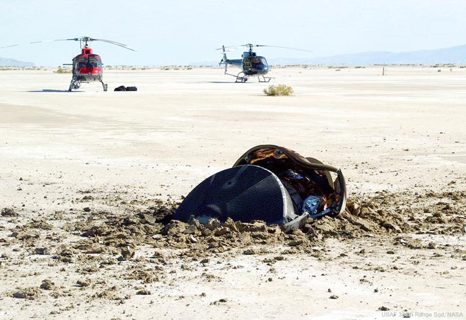 Das Bild zeigt die beschädigte Probenrückholkapsel der Raumsonde Genesis nach ihrem Absturz in der Wüste von Utah, im Hintergrund stehen zwei Hubschrauber.