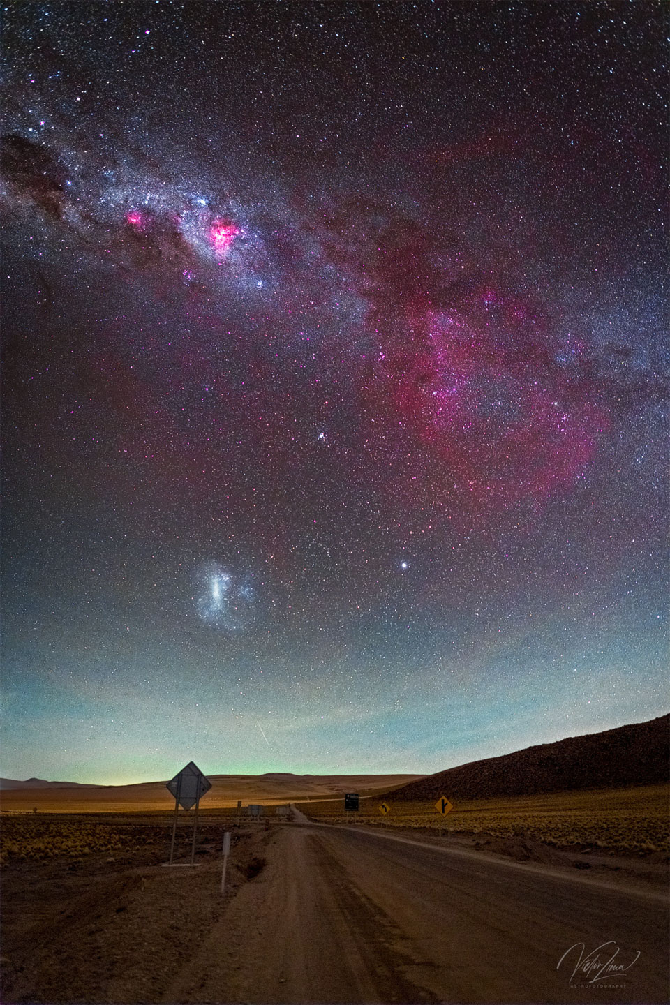 Das Bild zeigt eine großartige Himmelslandschaft mit einer braunen Wüstenstraße im Vordergrund und einen Himmel mit das galaktische Band der Milchstraße mit einem großen roten rechts, dem schwachen Gum-Nebel. Auch die Große Magellansche Wolke ist sichtbar.