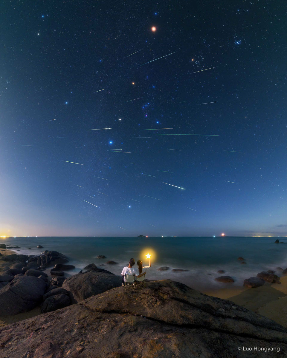 Das Kompositbild zeigt viele Meteorspuren die über einen Himmel mit dem bekannten Sternbild Orion ziehen. Im Vordergrund sitzen zwei Personen auf nebeneinander stehenden Stühlen, einer von ihnen hält einen Stab mit einem leuchtenden Stern am Ende.