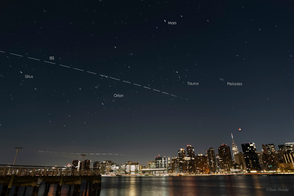 Das Bild zeigt eine Mondfinsternis über Manhattan, am Himmel sieht man die Plejaden, das Sternbild Orion und die Leuchtspur der Internationalen Raumstation.
