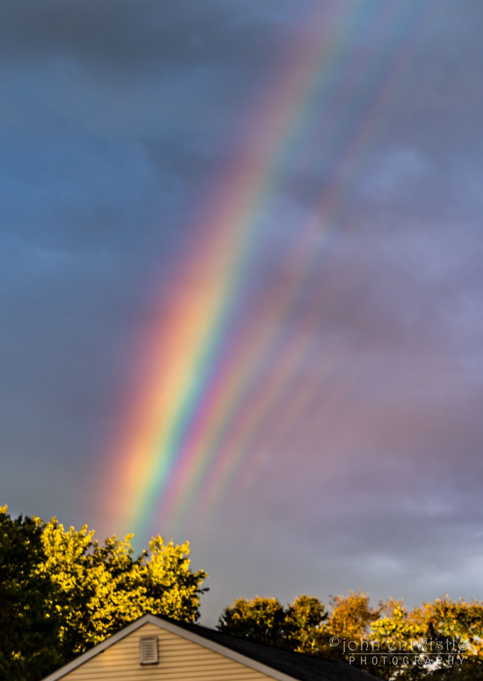 Das Bild zeigt einen Regenbogen mit einem markanten Interferenzmuster, das nur entsteht, wenn ale Regentröpfchen fast gleich groß sind. Im Vordergrund sind ein Dachgiebel und Bäume zu sehen.