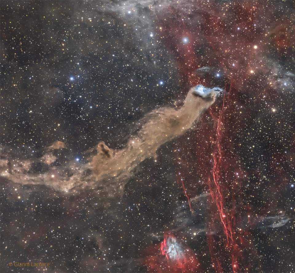 Der Reflexionsnebel im Bild ist eine dunkle längliche Form, deren Spitze blau leuchtet. Im Hintergrund sieht man rote Emissionsnebel einer früheren Supernovaexplosion.
