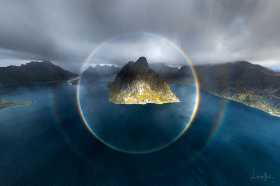 Das Bild zeigt einen kreisförmigen Regenbogen vor der Inselgruppe Lofoten, er wurde von einer Drohne vor den Inseln im Meer fotografiert.