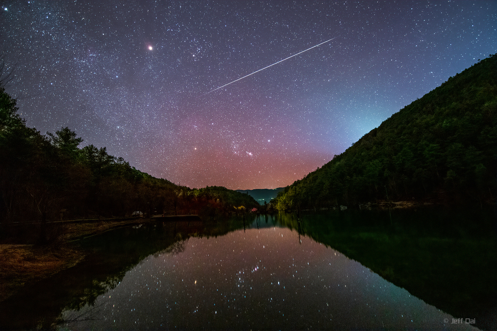 Das Bild zeigt einen ruhigen See im Tal des Blauen Mondes in der chinesischen Provinz Yunnan, in dem sich ein sternklarer Himmel mit dem Sternbild Orion und einem Meteor der Geminiden spiegelt.