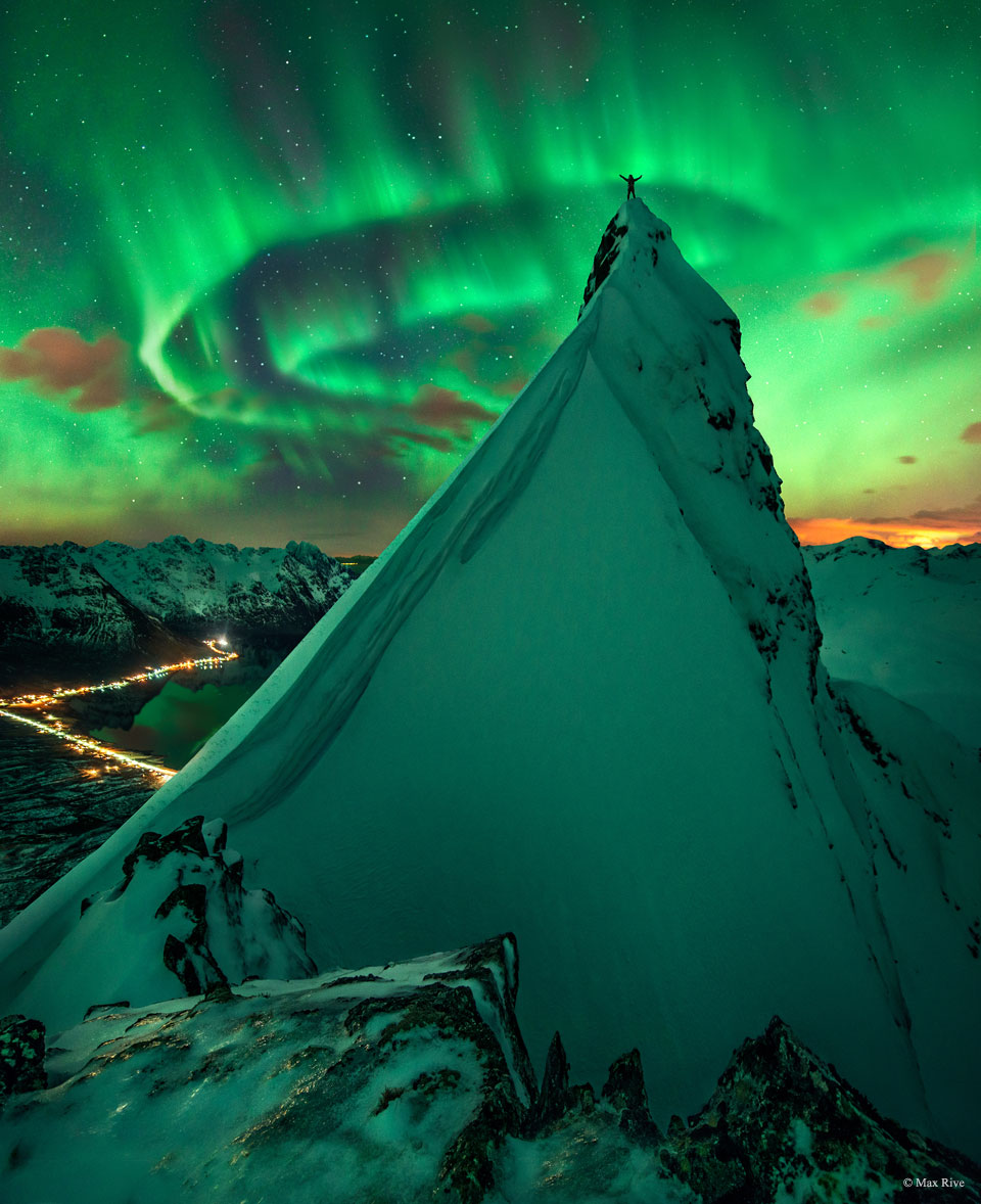 Auf einem steilen, schneebedeckten Gipfel steht eine Person mit erhobenen Armen, im Hintergrund leuchtet am Himmel ein helles Polarlicht.