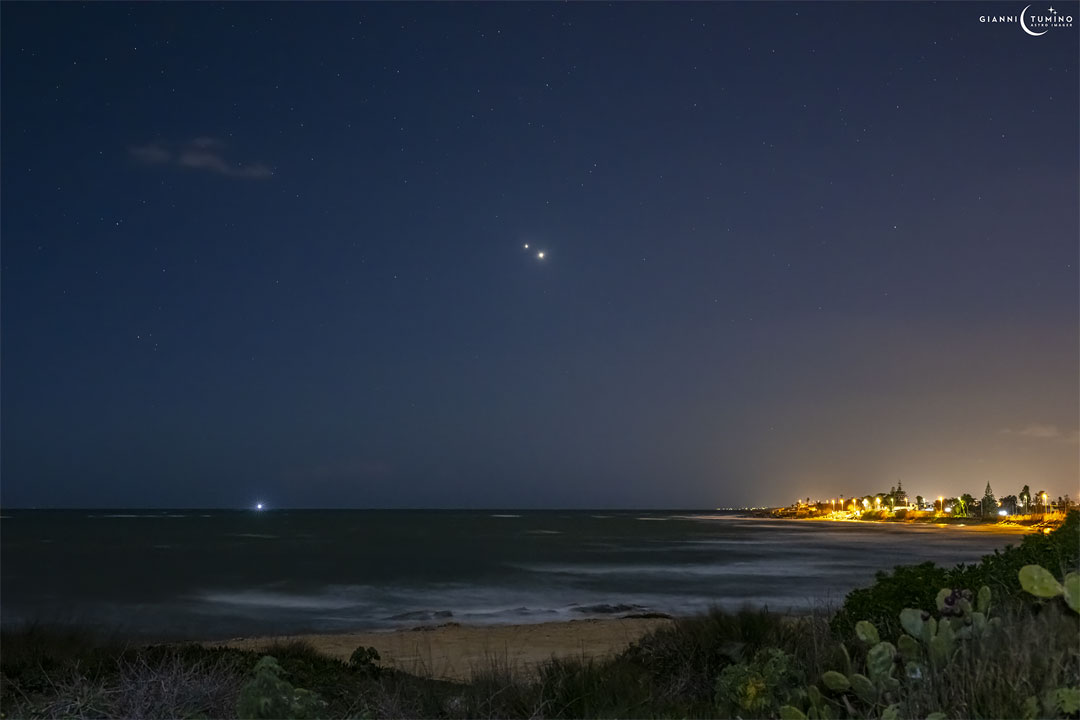 Zwei helle Objekte stehen am Nachthimmel voller Sterne sehr nahe beieinander. Im Vordergrund ist ein Strand zu sehen, an dem einige beleuchtete Strukturen über dem Wasser zu sehen sind.