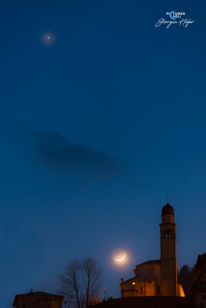 Neben der dunklen Silhouette einer Kirche leuchtet eine schmale Mondsichel mit Erdlicht, hoch darüber leuchtet oben der Planet Venus.