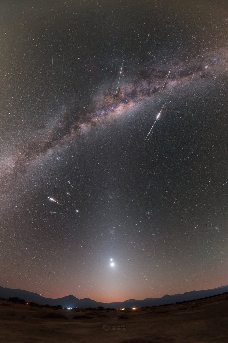 Über einem gebirgigen Horizont leuchten zwei helle Planeten, darüber wölbt sich am sternklaren Himmel die Milchstraße mit markanten Dunkelwolken. Das Bild ist von teilweise hell aufblitzenden Meteorspuren durchzogen, die von einem gemeinsamen Punkt ausströmen.
