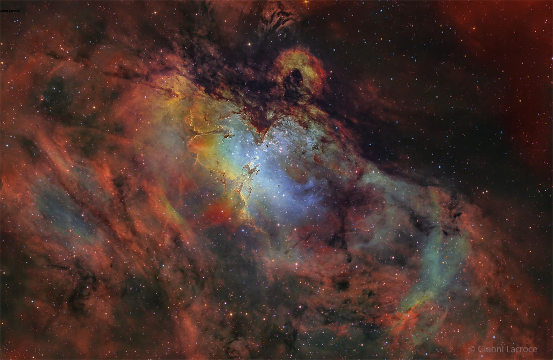 Die Säulen der Sternbildung in der Mitte vor blau leuchtendem Hintergrund ist von roten, orangefarbenen und dunklen Nebeln umgeben. 