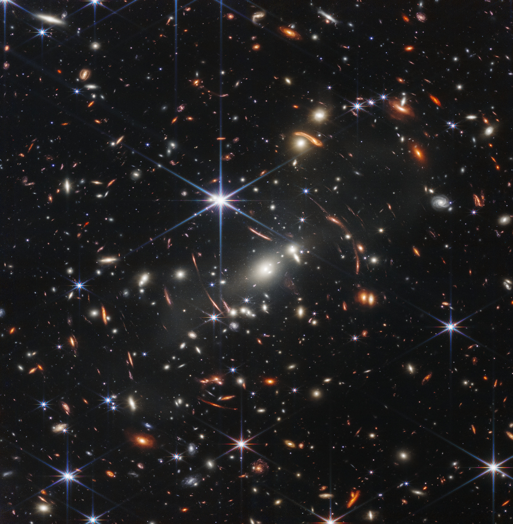 Das Bild ist von hellen leuchtenden Objekten übersät, von denen die meisten Galaxien sind. In der Mitte leuchtet ein Stern mit den für das JWST typischen Zacken.
